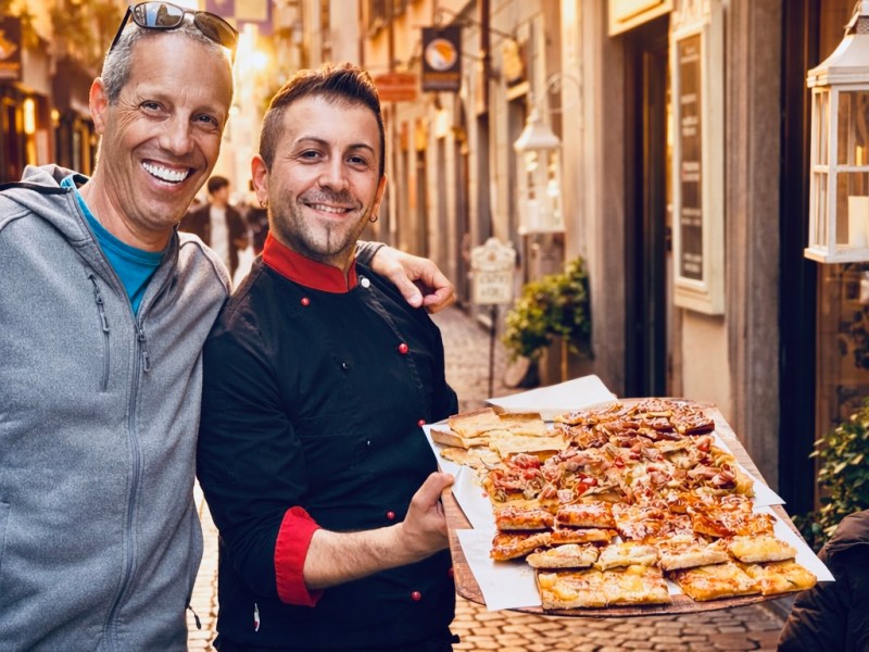 Two men posing with a tray of assorted pizzas on a sunlit cobblestone street.