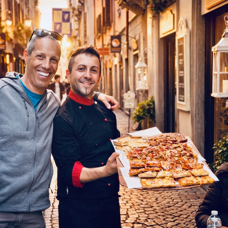 Two men posing with a tray of assorted pizzas on a sunlit cobblestone street.