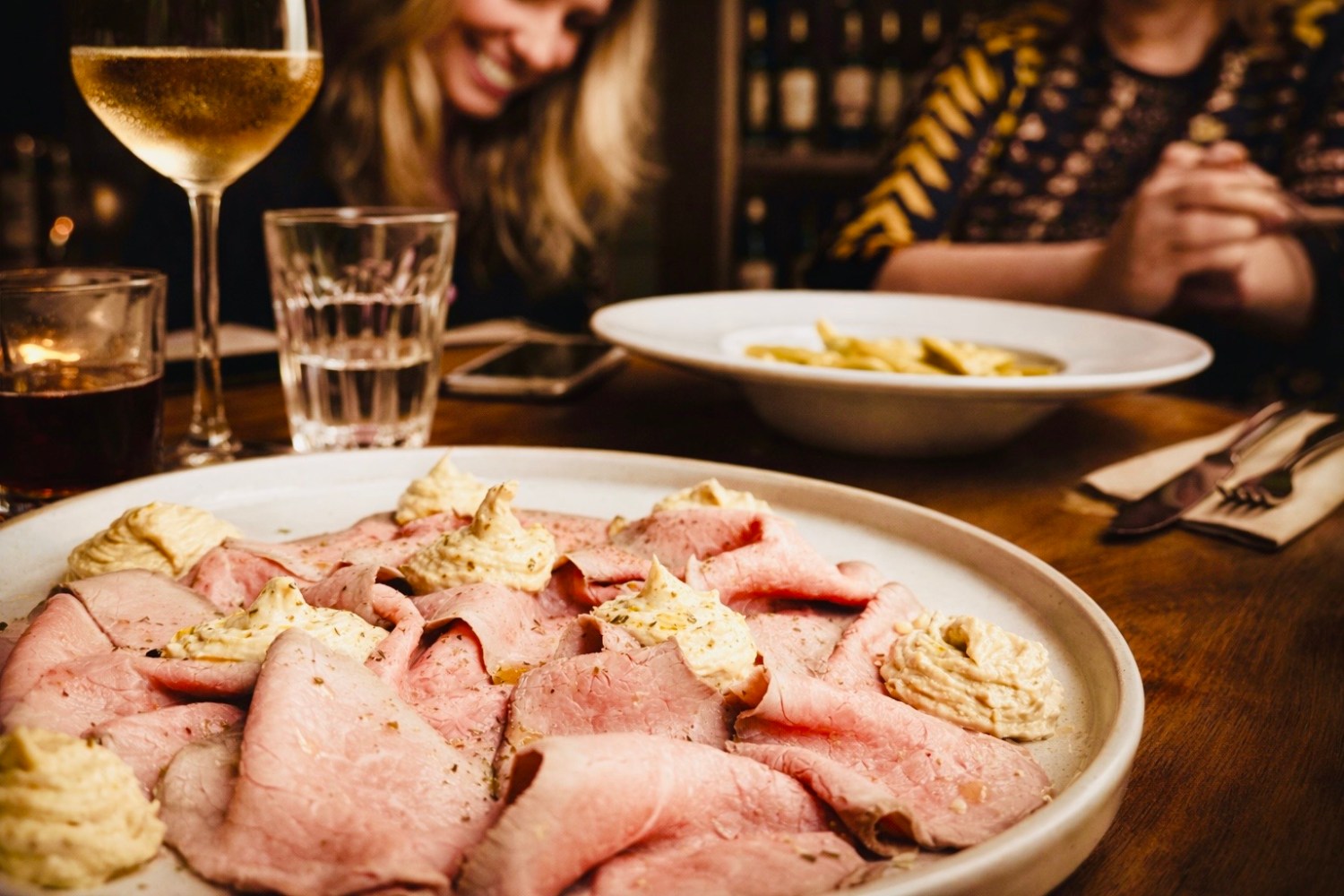 Plate of sliced roast beef with sauce on a table, people blurred in background.