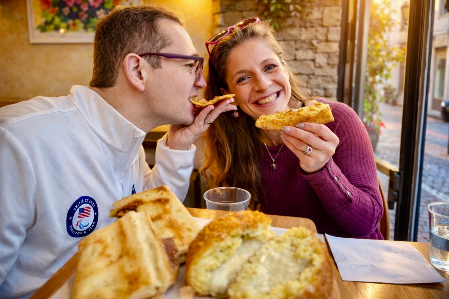 Smiling couple sharing sandwiches at a cozy cafe table with sunlight streaming in.