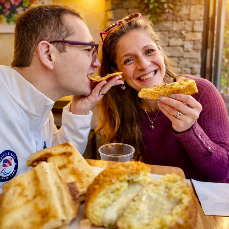 Smiling couple sharing sandwiches at a cozy cafe table with sunlight streaming in.