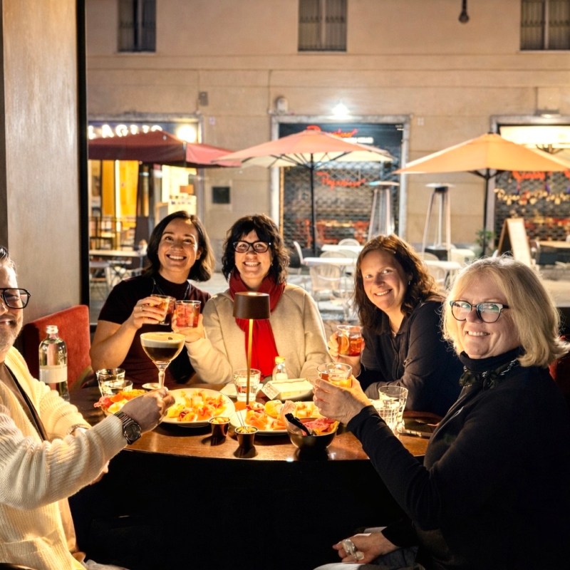 Five people smiling at a restaurant table with drinks and snacks inside a warmly lit dining area.