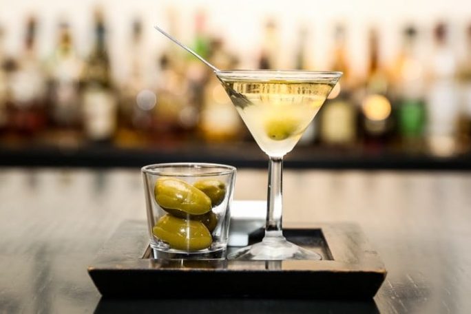 Martini glass with olive and a glass of olives on a tray in a bar setting.