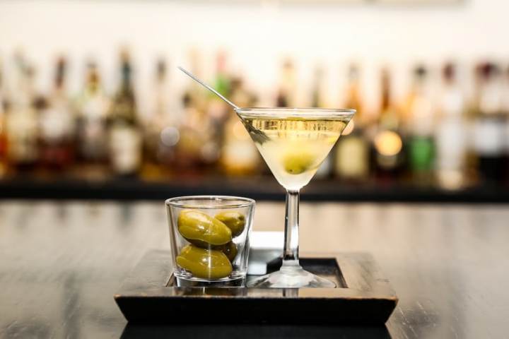 Martini glass with olive and a glass of olives on a tray in a bar setting.
