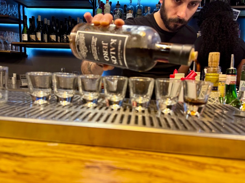 Bartender pouring shots from a large bottle into seven shot glasses on a bar counter.