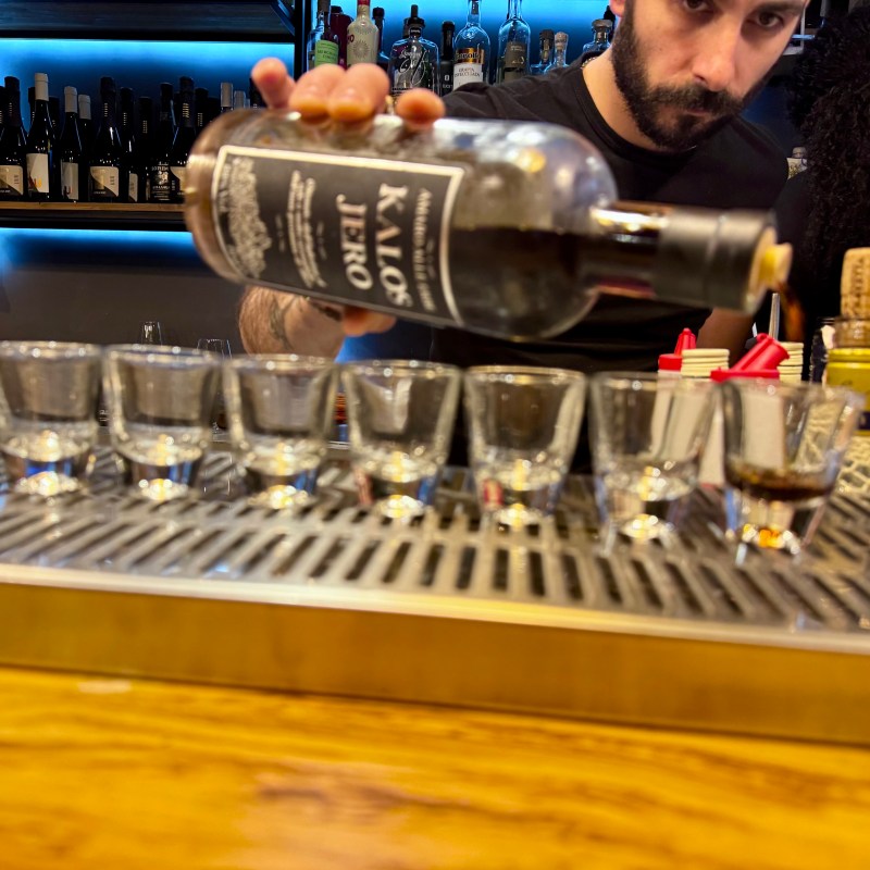 Bartender pouring shots from a large bottle into seven shot glasses on a bar counter.