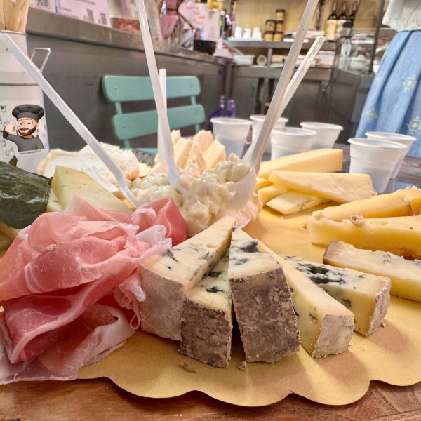 A wooden board with assorted cheeses and prosciutto slices, garnished with leaves and skewers.