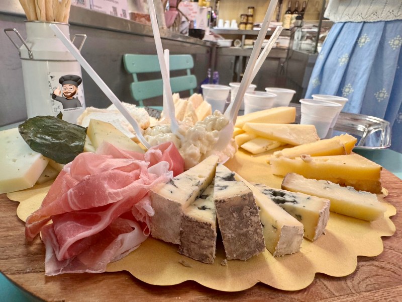 A wooden board with assorted cheeses and prosciutto slices, garnished with leaves and skewers.