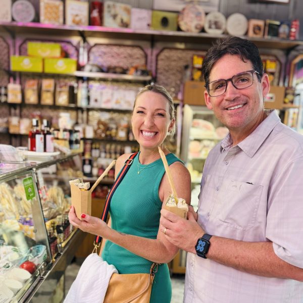 Two people smiling and holding food cups in a deli with various products displayed.