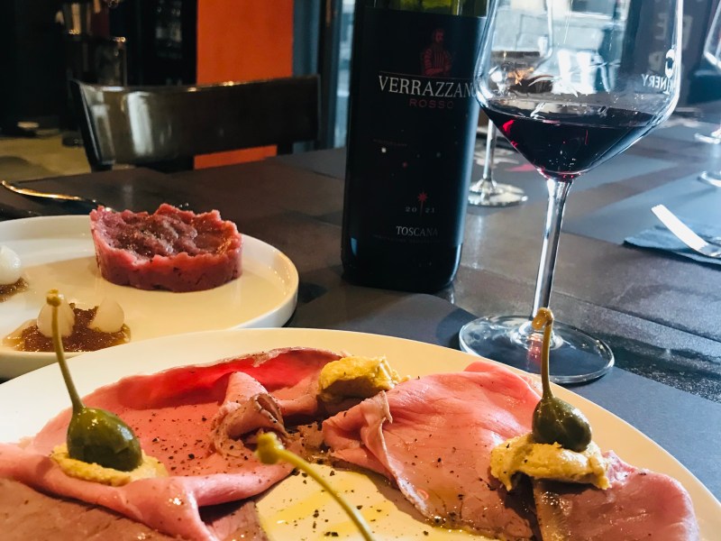 Sliced meat with capers and mustard on a plate next to a wine glass and bottle on a table.
