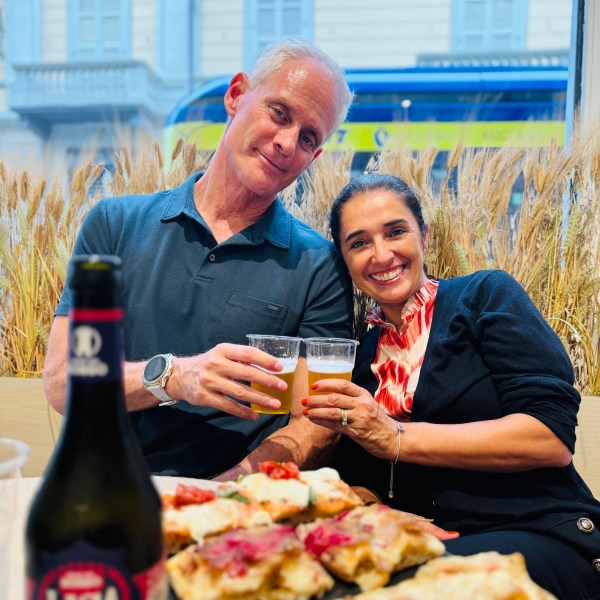 Two people smiling, toasting with beer, with pizza and a beer bottle on a table in front of them.