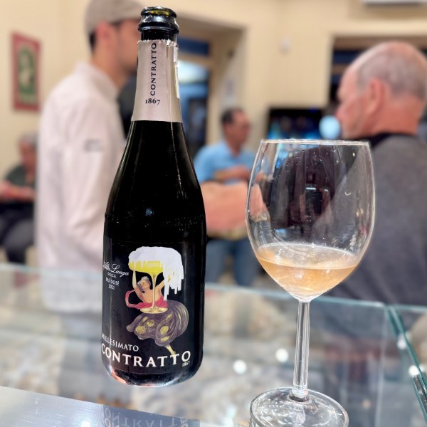 Bottle of Contratto wine and a glass on a glass counter, people in background.