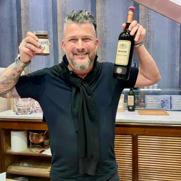 Person smiling, holding a wine bottle in one hand and a jar in the other, standing in a kitchen.