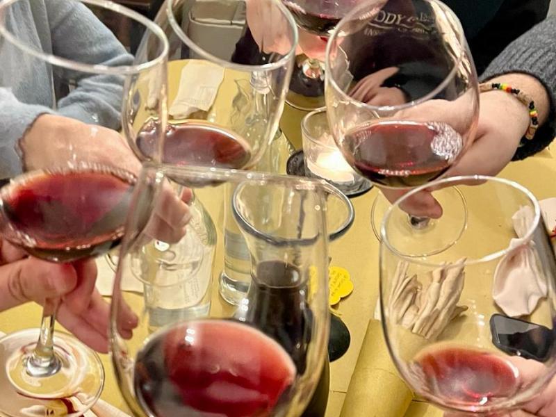 Six people toasting with red wine glasses at a table with yellow tablecloth.