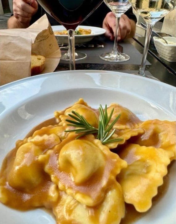 Ravioli with sauce and rosemary garnish on a plate with wine glasses in the background.