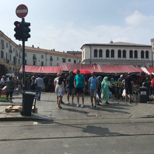 Busy outdoor market with people, stalls, and a red-striped canopy on a sunny day.