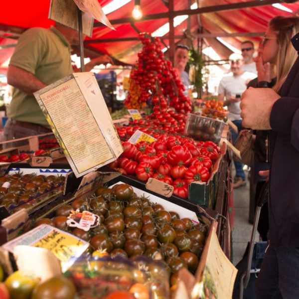 People browsing tomatoes at an outdoor market stall with red umbrellas.