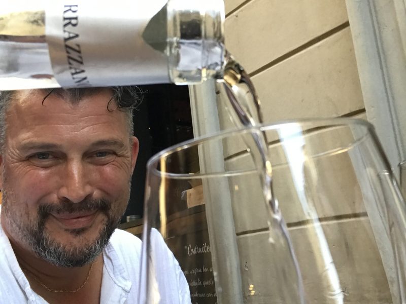 Man in white shirt smiling while pouring wine into a glass outdoors.