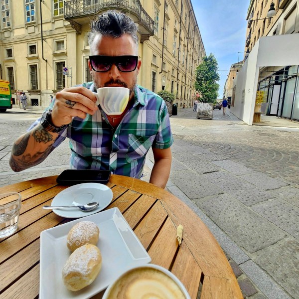 Person with sunglasses drinks coffee at an outdoor cafe table with pastries and cappuccino.