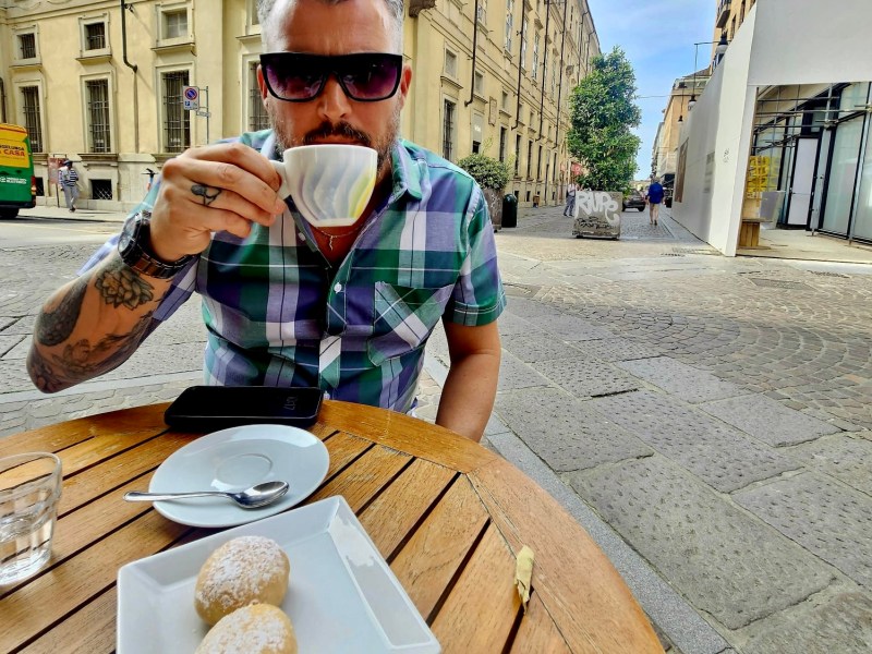 Person with sunglasses drinks coffee at an outdoor cafe table with pastries and cappuccino.