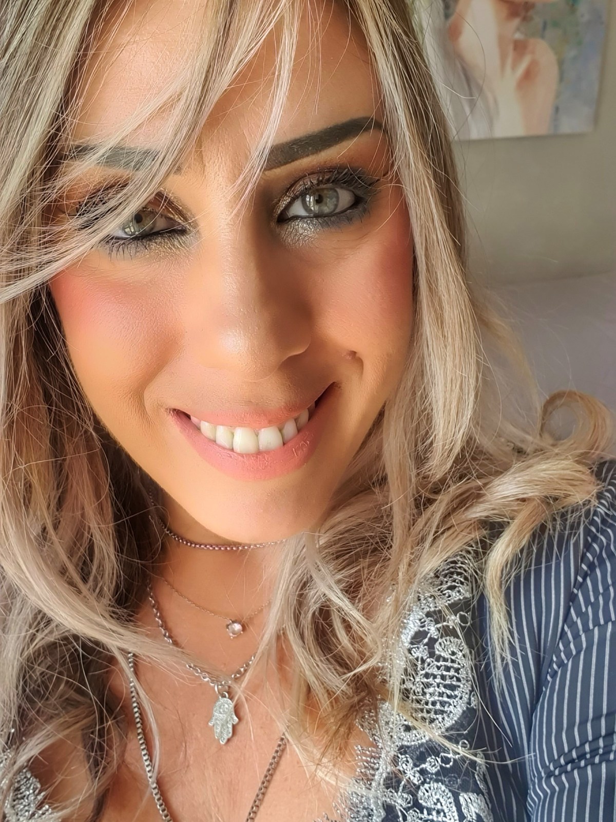 Woman smiling with blonde hair and a necklace, artwork in background.