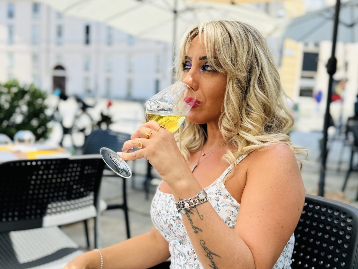 Woman with blonde hair drinking from a glass under an outdoor umbrella.