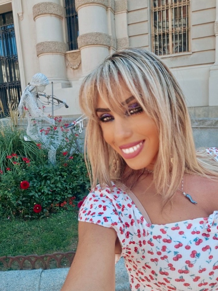 Person with blonde hair and cherry-patterned dress taking a selfie near sculpture and building.