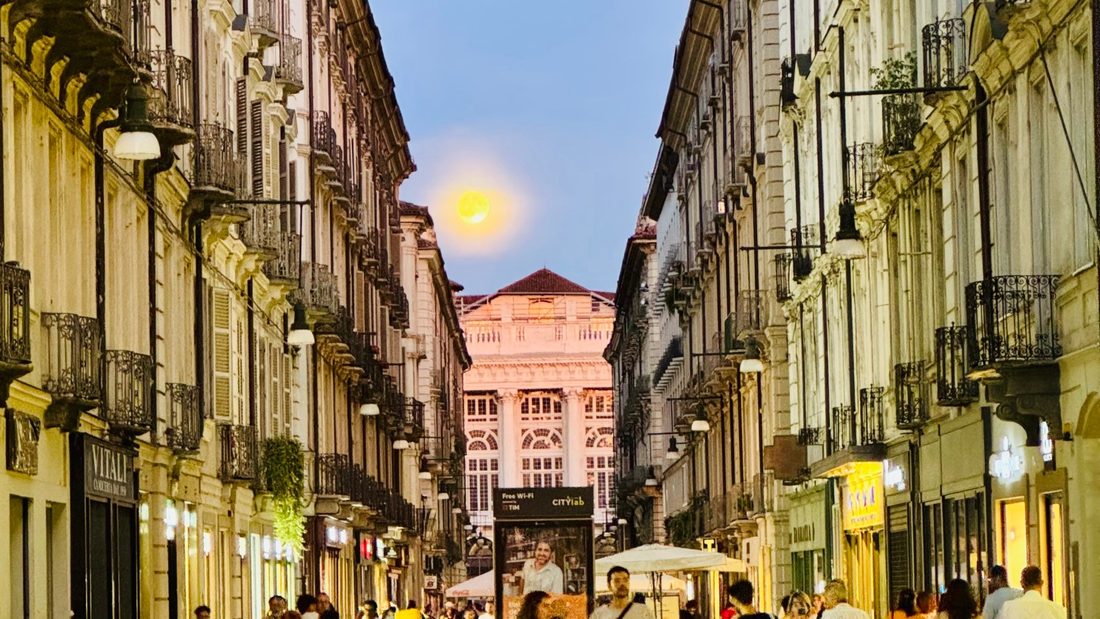 People walking on a narrow street with historic buildings under a rising moon.