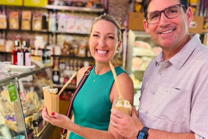 Two people smiling, holding food in a shop with shelves of products in the background.
