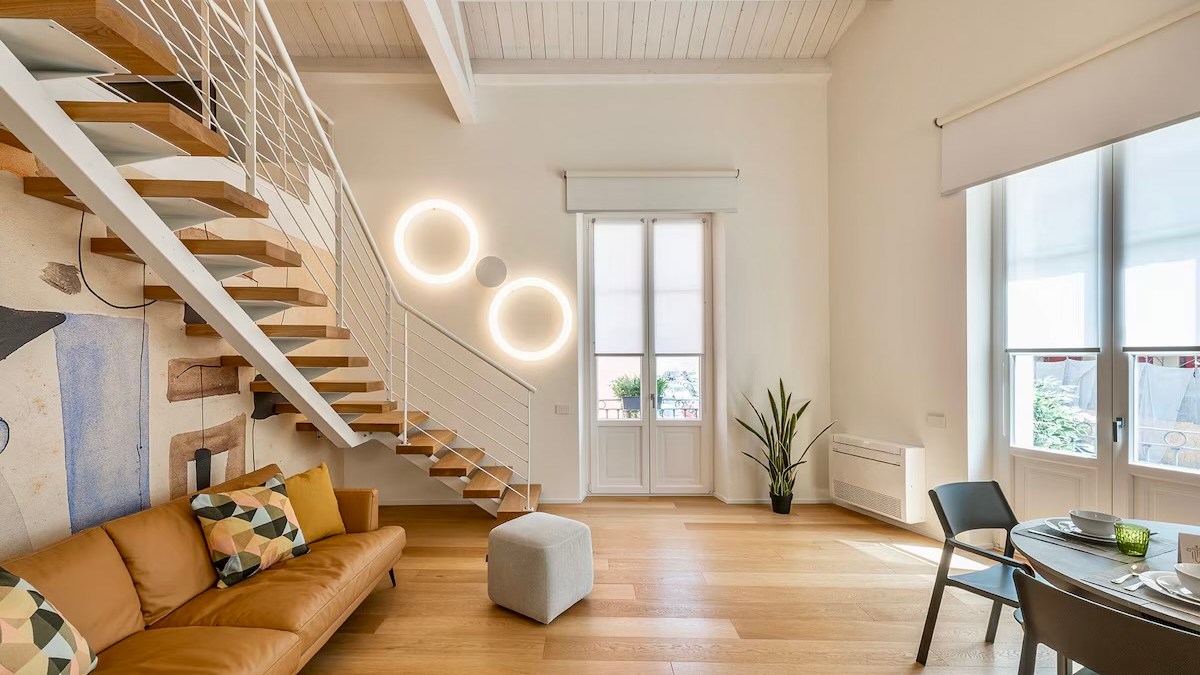 Modern living room with staircase, sofa, plant, and dining table near large windows.
