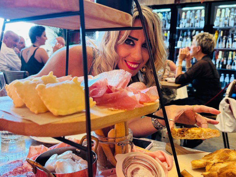 Woman smiling at table with cheese, meats, and bread in restaurant setting.