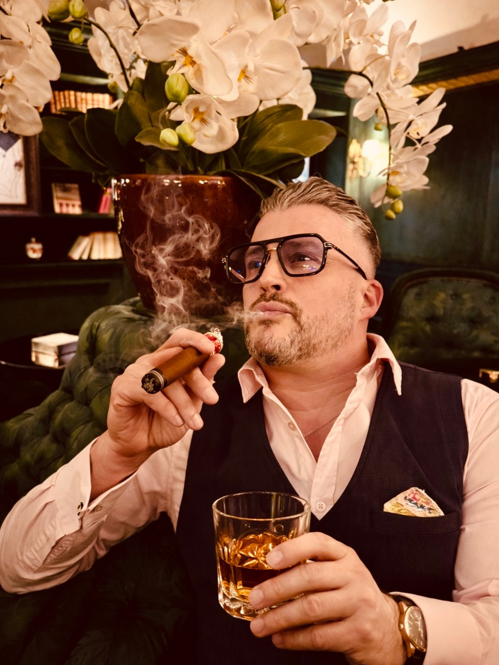 Man with sunglasses smokes a cigar and holds a drink, sitting near white orchids.