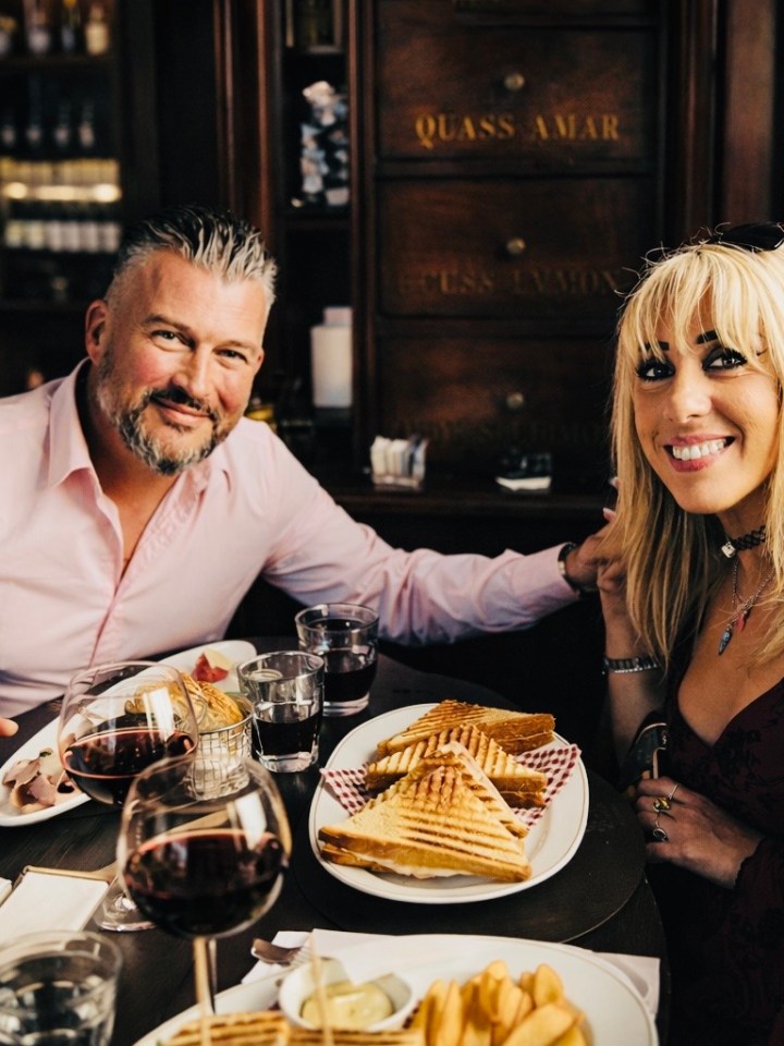 A smiling couple dining at a restaurant with wine, sandwiches, and fries on the table.