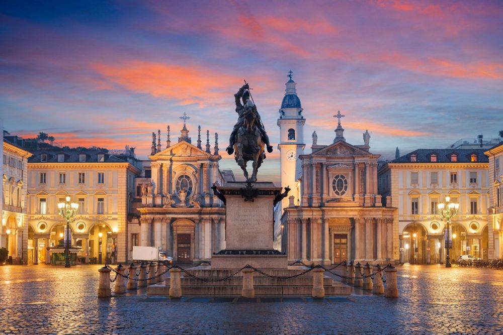Equestrian statue in a square at sunset, framed by historic buildings and a colorful sky.