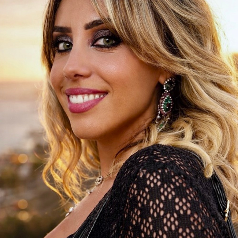 Smiling woman with wavy hair and large earrings against a sunset background.