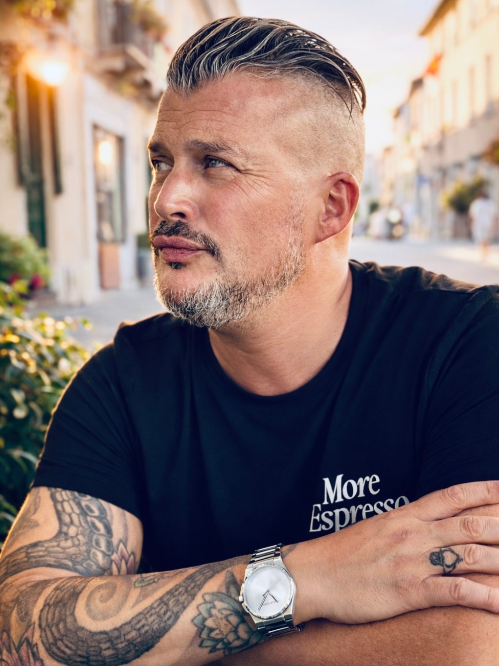 Man with tattoos and watch, sitting in street, wearing a T-shirt with 'More Espresso' text.