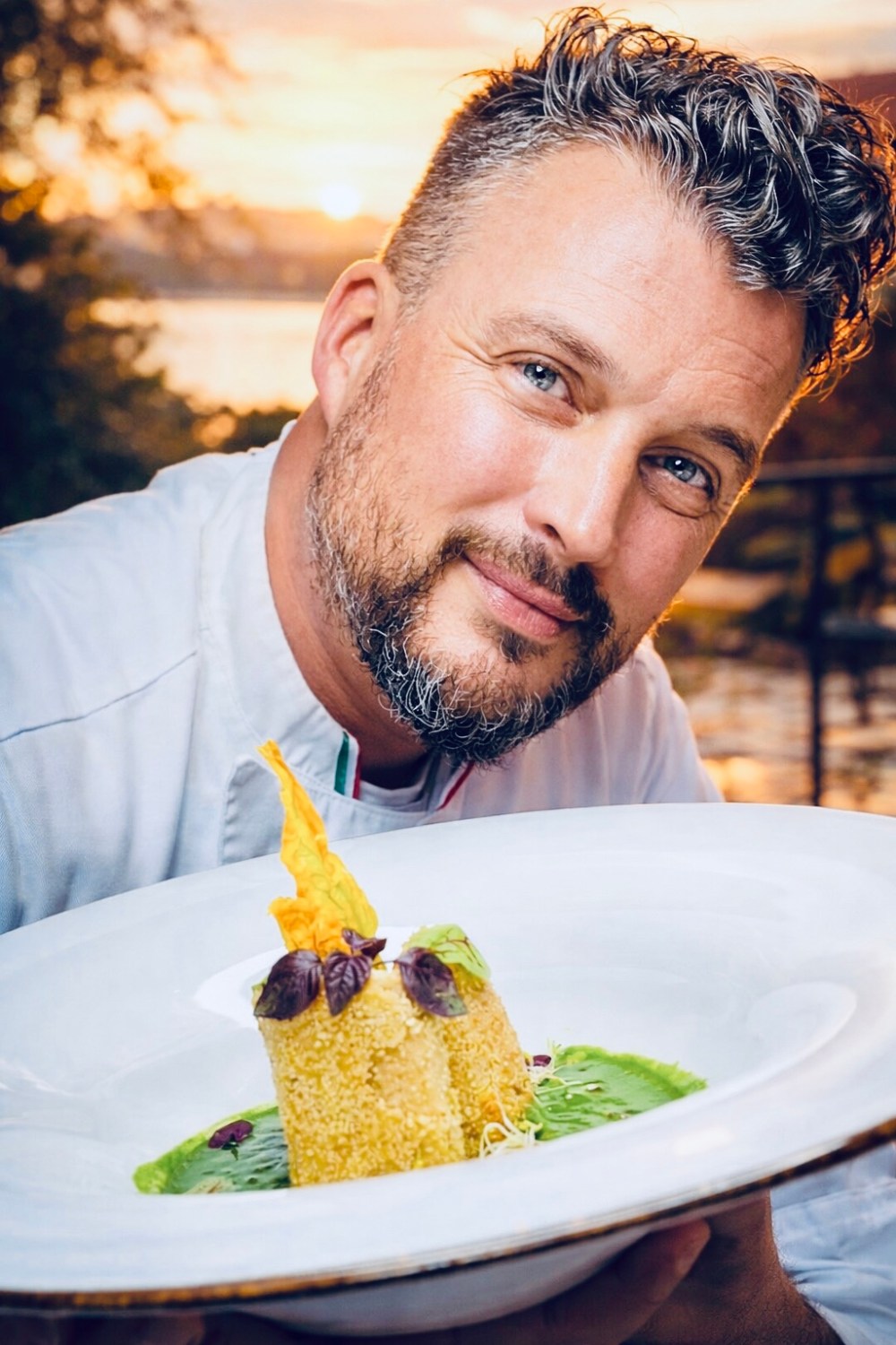 Chef holding a plate with a gourmet dish against a sunset backdrop.