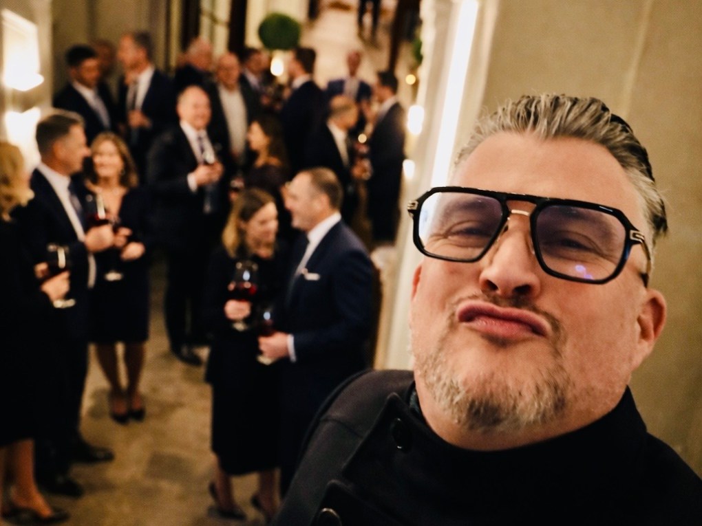 Man in glasses taking a selfie at a formal gathering in a hallway.