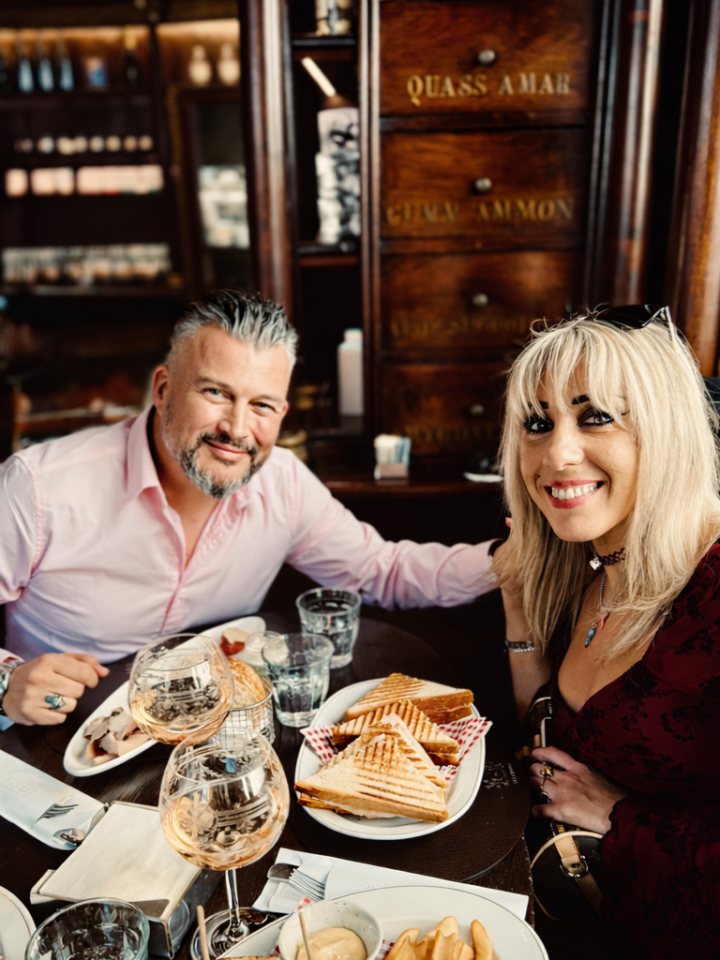 Two people dining at a restaurant smiling with plates of sandwiches and drinks on the table.