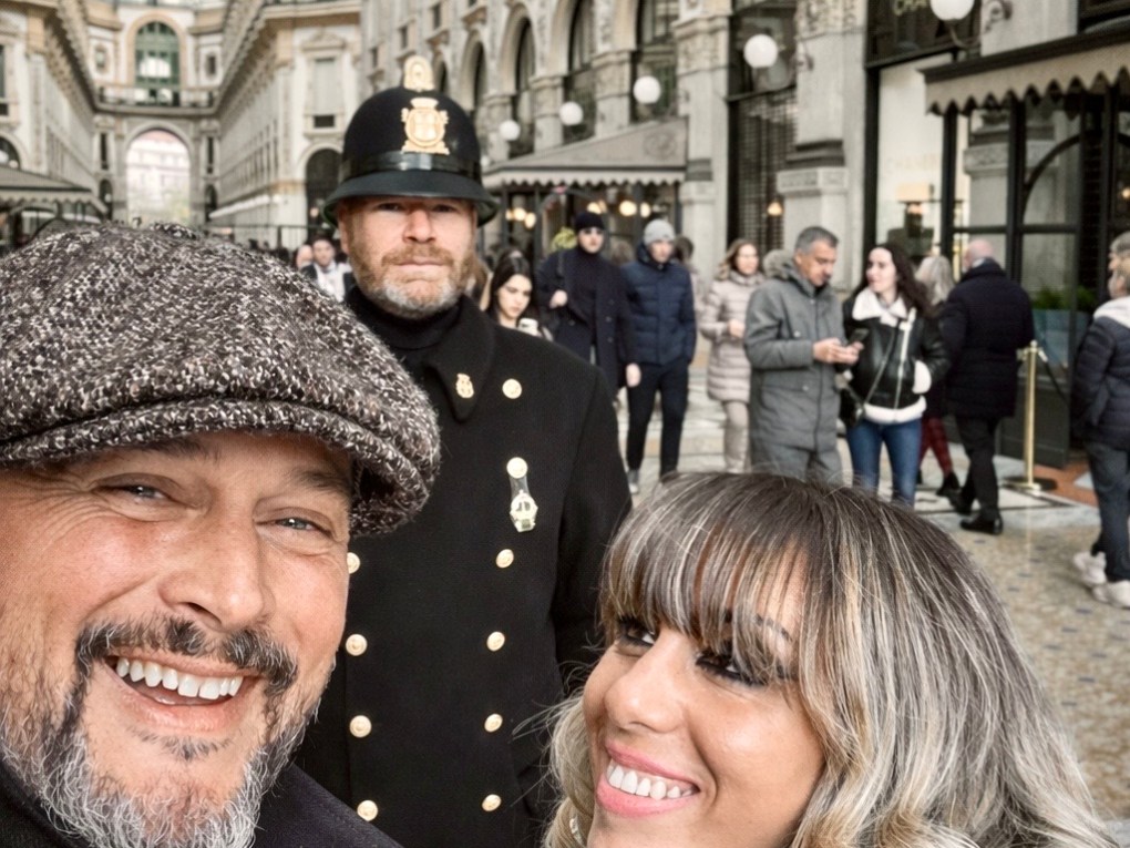 Smiling couple takes a selfie in an ornate shopping arcade, with a uniformed officer standing behind.