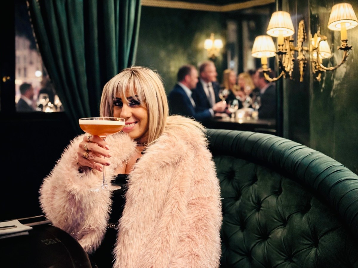 Woman in a pink fur coat holding a cocktail in a dimly lit restaurant.