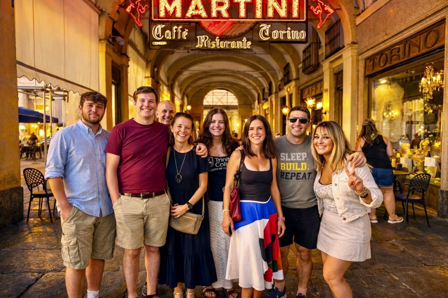 Group of eight people smiling under a lit 'Martini' sign at an outdoor cafe.