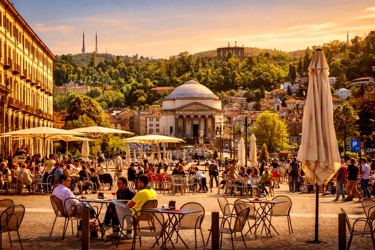 Outdoor café with people under umbrellas, buildings, and lush hills in the background at sunset.