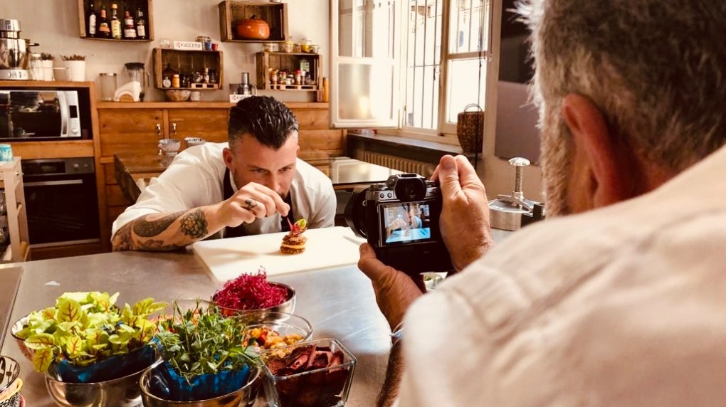 Chef garnishing food while man photographs in a rustic kitchen.