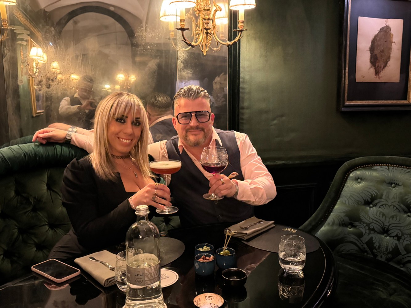 Two people smiling and toasting drinks at a dimly lit restaurant table.