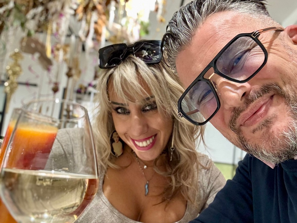 Two people smiling, sitting at a table with drinks and food, indoors with decorative background.