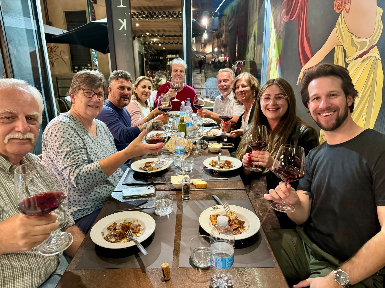 Group of people dining together, holding glasses of red wine, smiling at the camera.