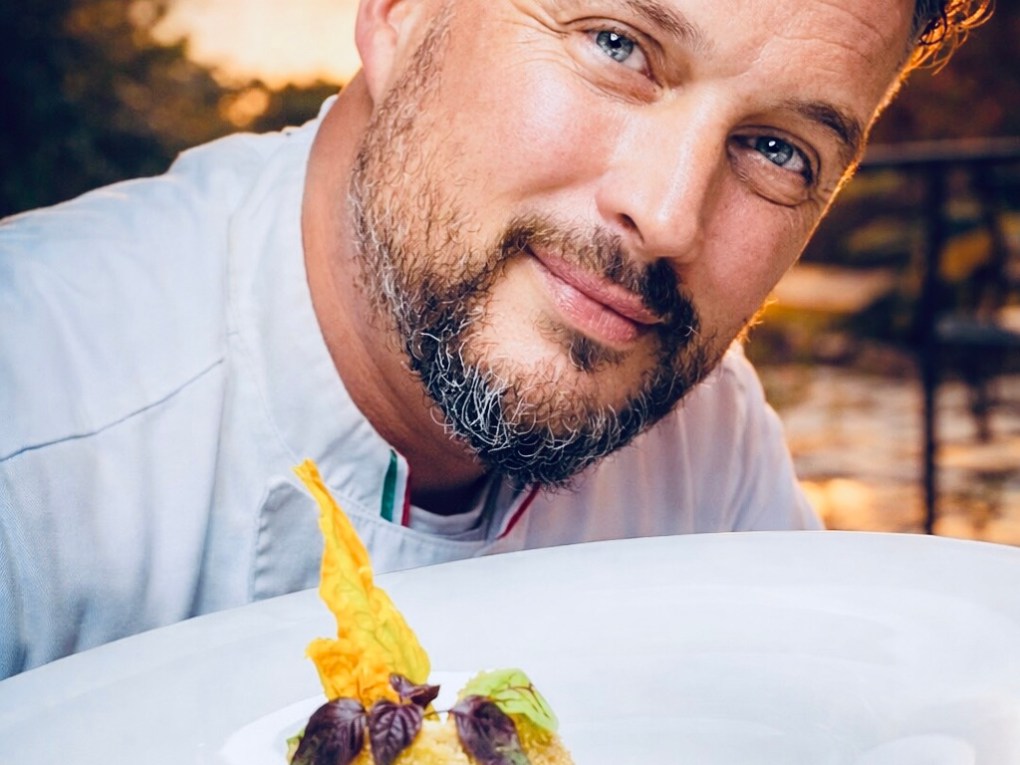 Smiling man holding a plate with gourmet food garnished with herbs outside at sunset.