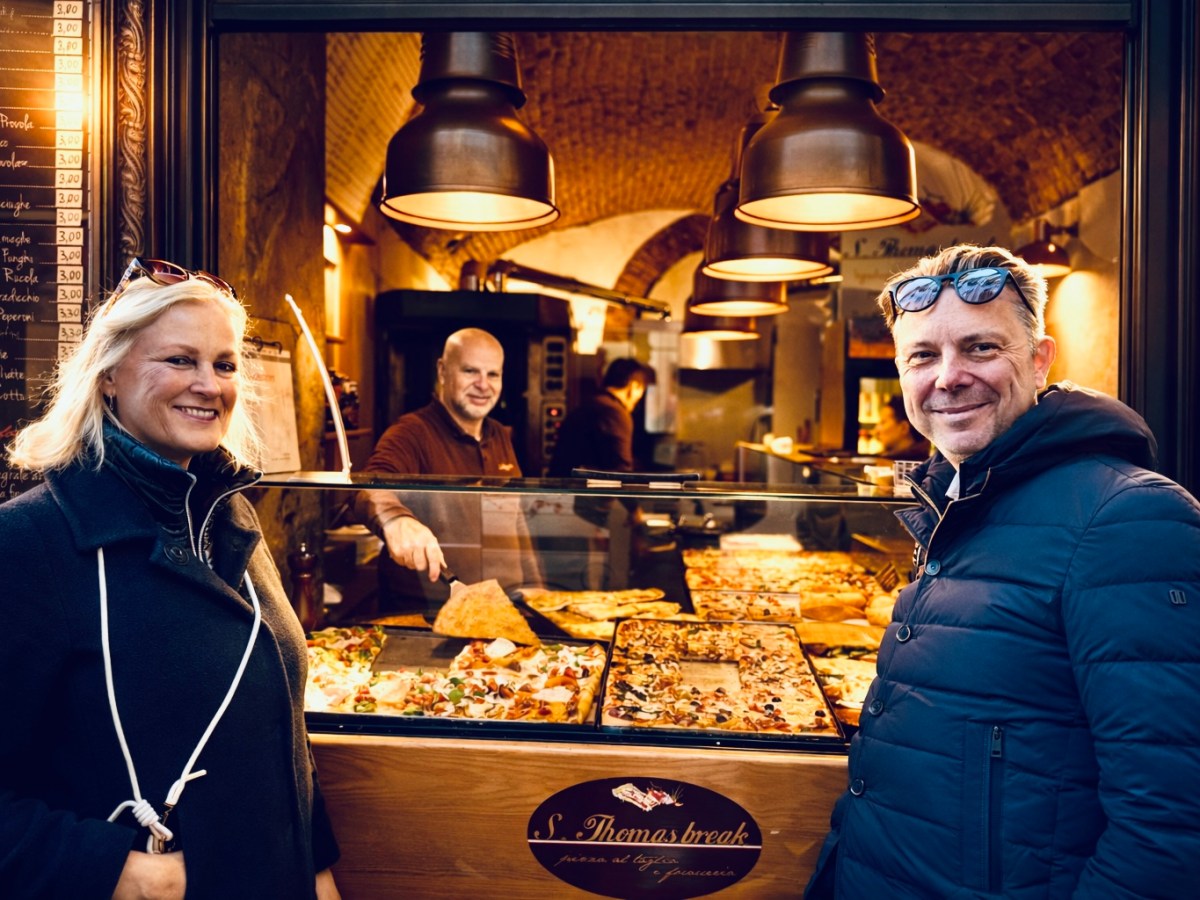 Two people smiling outside a pizzeria with pizza displayed behind the glass.