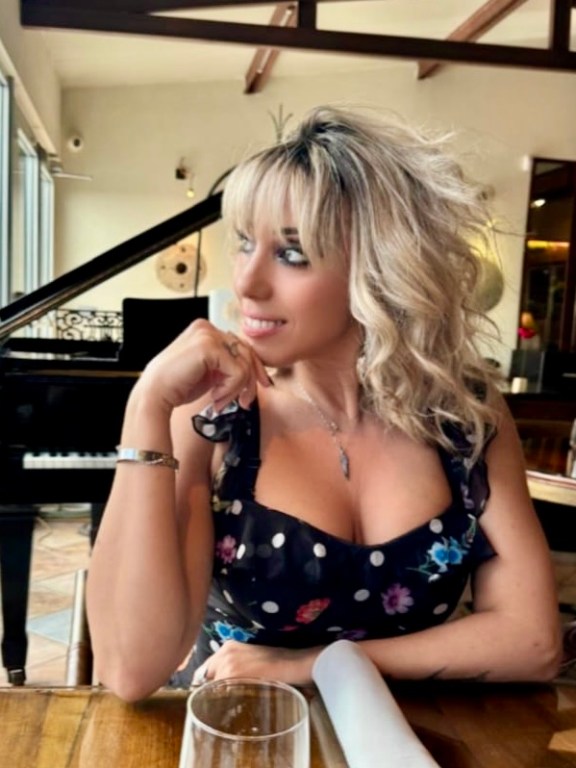 Woman with blonde hair in a black dress sits by a piano in a restaurant.